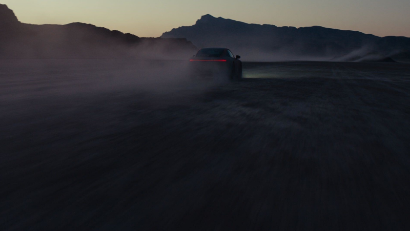 A vehicle silhouette emerges through desert dust at dawn, creating dramatic atmosphere in moody automotive cinematography against mountain backdrops.