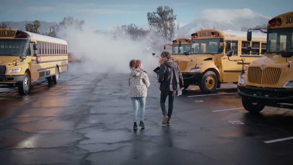 The director and the actress walking amidst a collection of parked yellow school buses, engaged in a discussion about the upcoming scene.