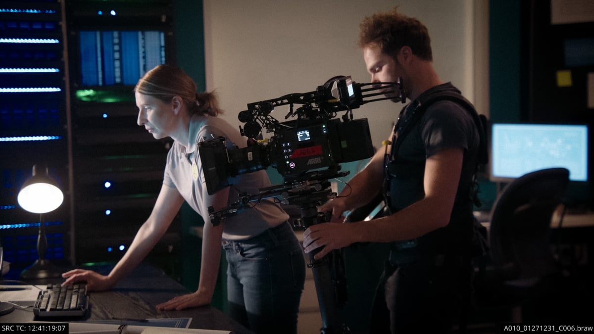 A cameraman wearing a strapped camera around his body, capturing a scene of the actress in front of her computer
