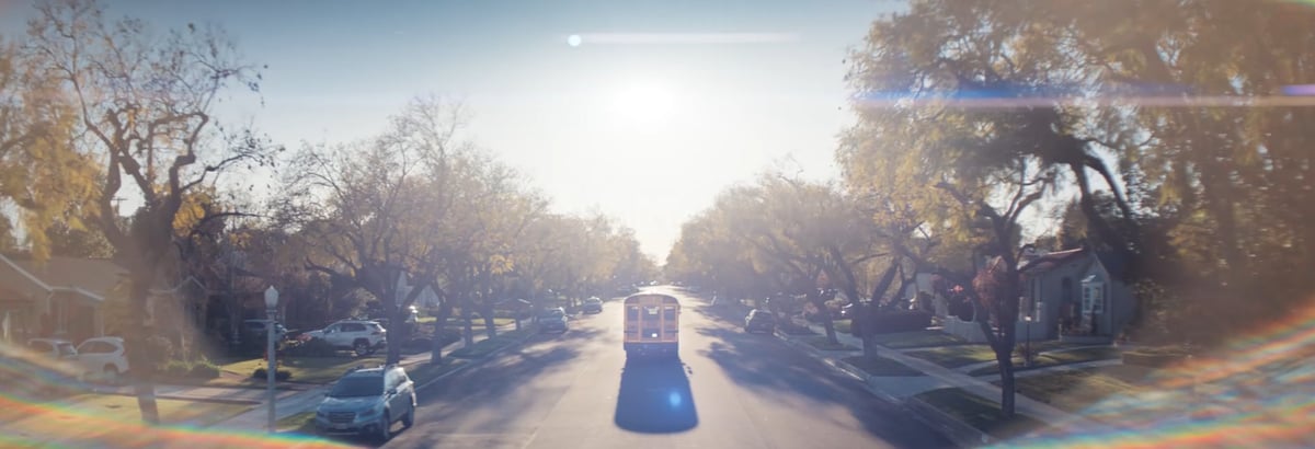 An aerial view capturing the rear of a school bus in motion on a road, surrounded by trees and basking in the gentle sunlight