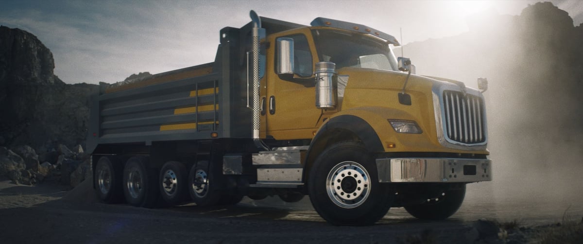 A side view of a sizable yellow truck, with sunlight behind it casting a reflective glow onto the front.