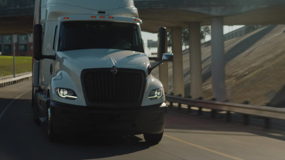 A white International semi-truck approaches head-on with its LED headlights illuminated as it passes under a highway overpass.