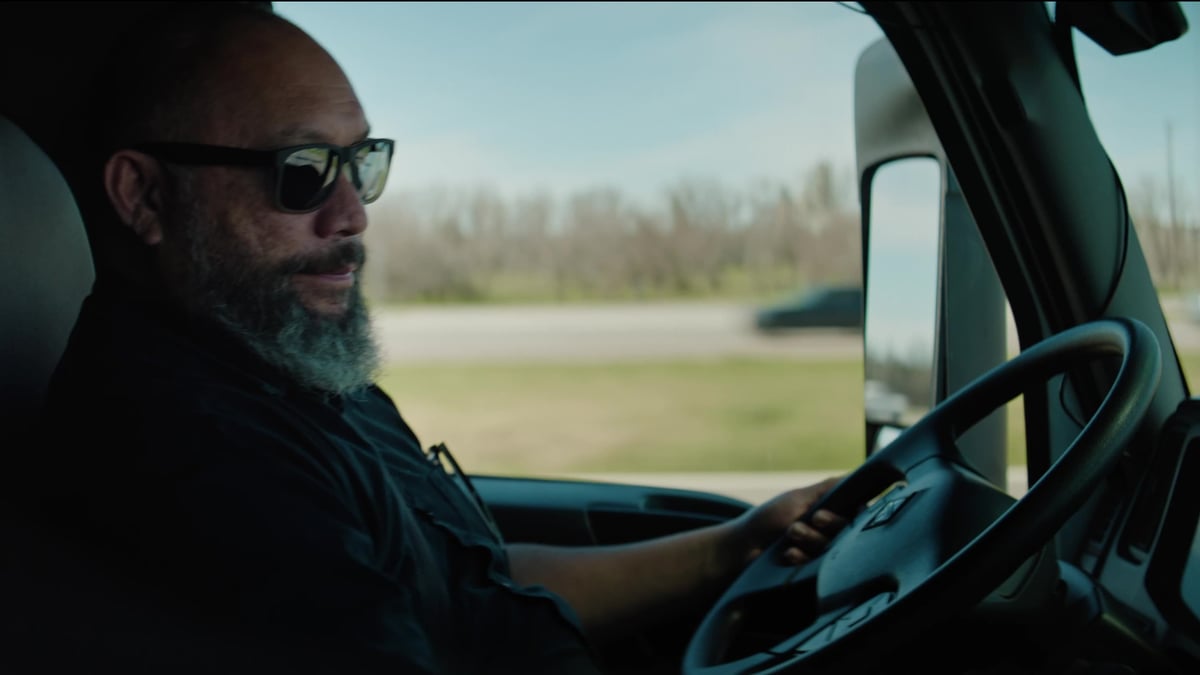 A bearded truck driver wearing sunglasses sits behind the wheel of a truck, gazing ahead at the open road.
