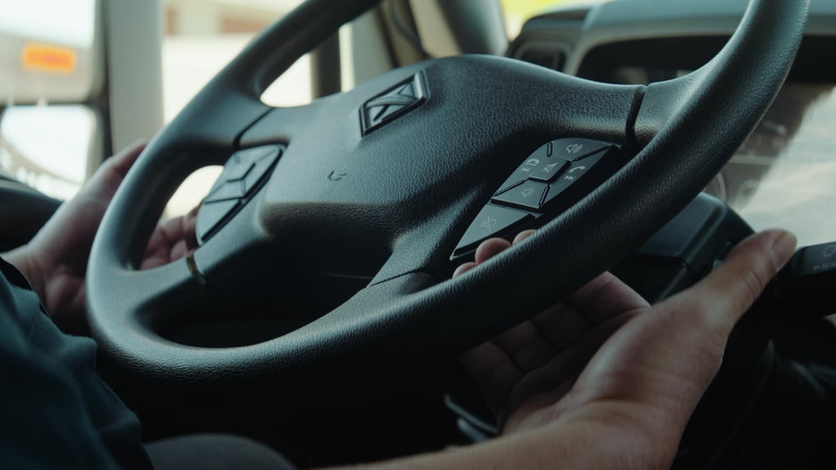 Two hands grip a truck's steering wheel, which features the International diamond logo and integrated control buttons.