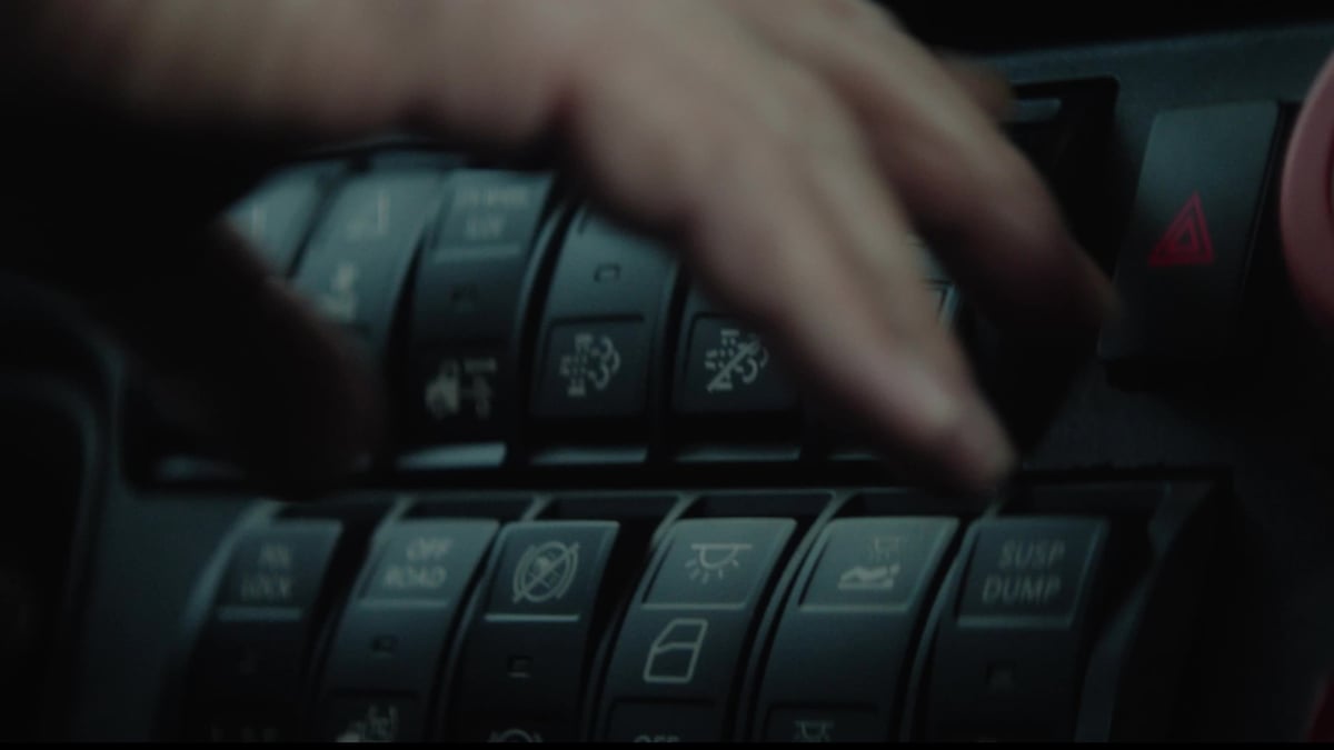 A hand reaches over a panel of illuminated control switches inside a truck cab in a dimly lit close-up shot.
