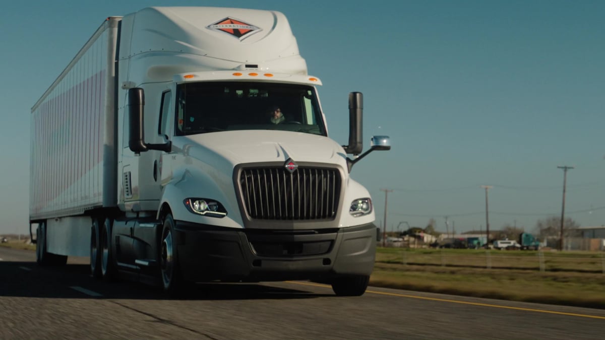 A white International semi-truck with a striped trailer approaches directly from the front on a rural highway under a clear blue sky.