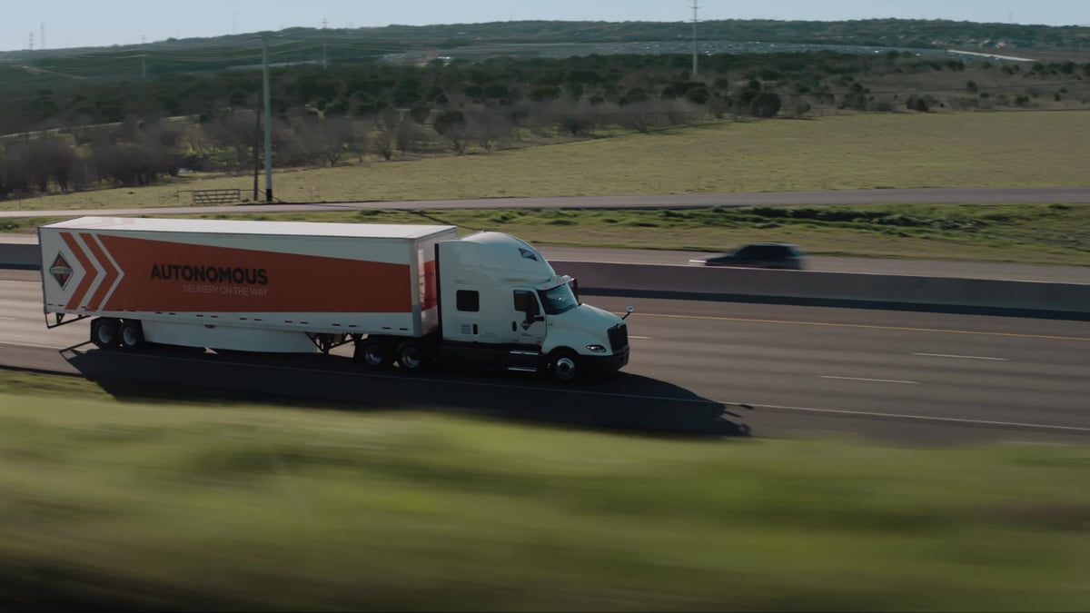 A white International semi-truck with a bold orange trailer reading "AUTONOMOUS — Delivery on the Way" travels along a Texas highway.