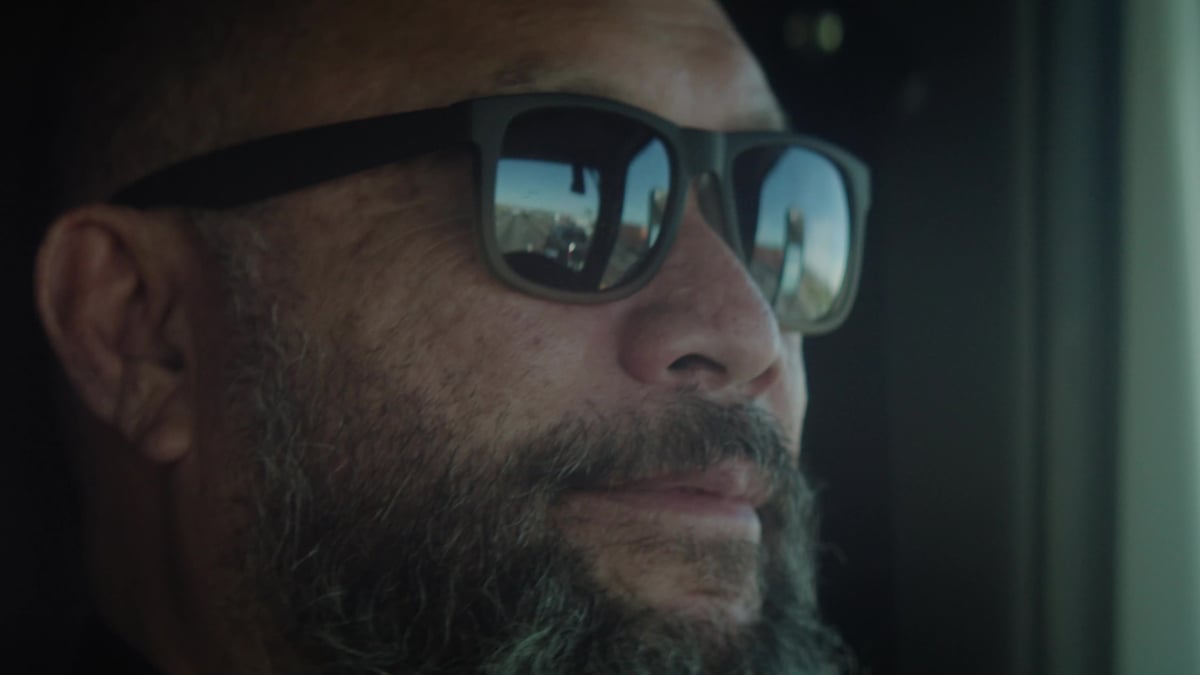 A close-up of a bearded truck driver's face shows the open road reflected in his sunglasses as he looks ahead.