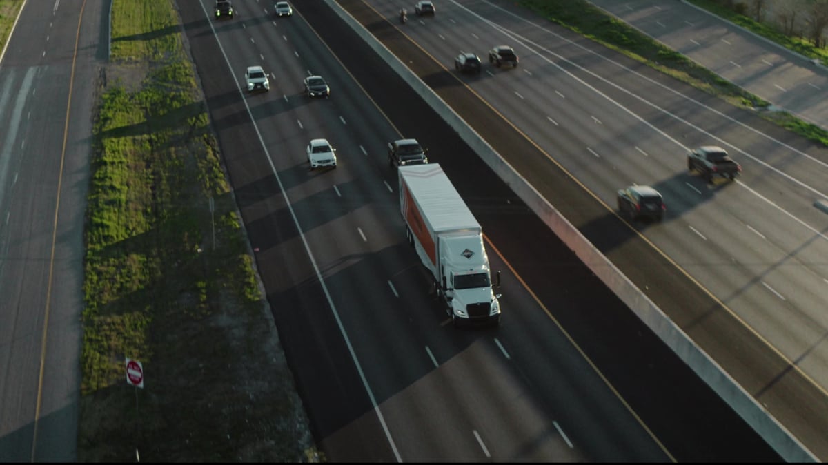 An aerial drone shot shows a white semi-truck with an orange trailer traveling among highway traffic at golden hour.