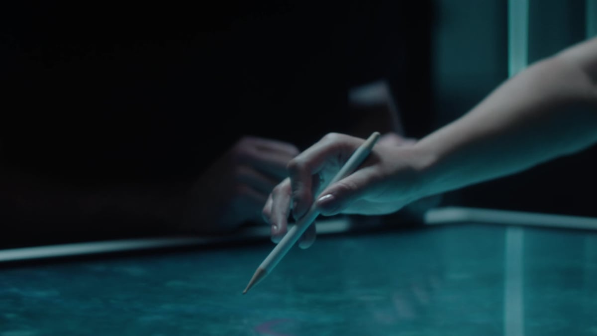 A hand holds a stylus or pencil over a glowing teal surface, with another hand visible in the blurred background.