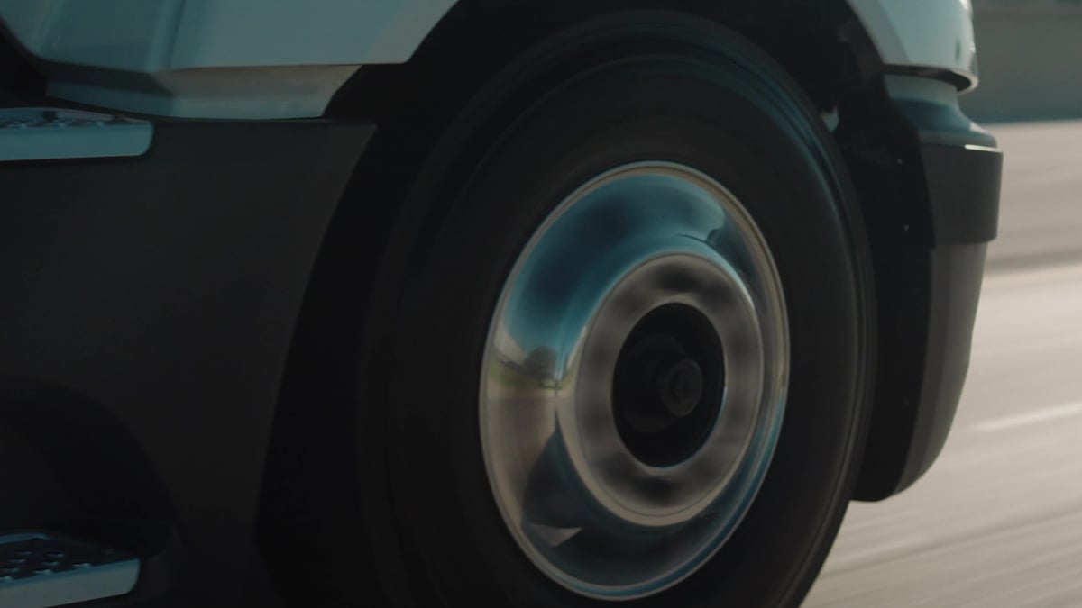 A close-up of a spinning truck wheel and tire captures motion blur against a blurred road surface and light blue truck body.