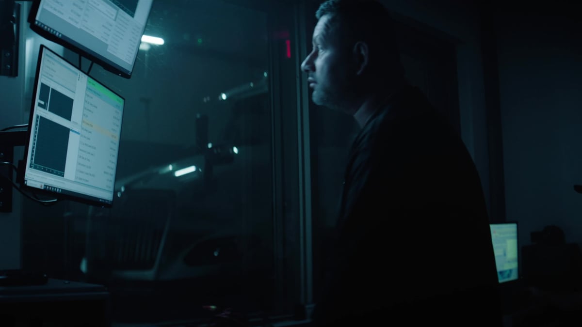 A man sits in profile in a dark room, his face illuminated by the glow of multiple computer monitors displaying data and spreadsheets.