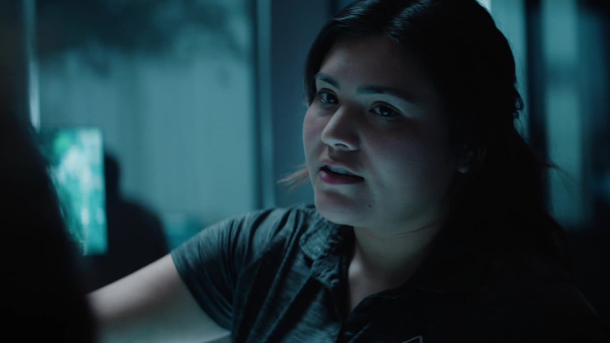 A young woman with dark hair looks forward with an urgent or concerned expression in a dimly lit setting.