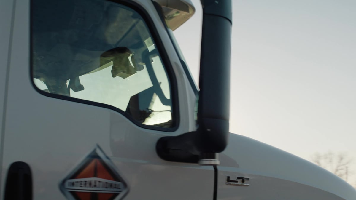 A low-angle exterior shot of an International LT semi-truck cab shows the driver's window, side mirror, and branded logo against a pale sky.