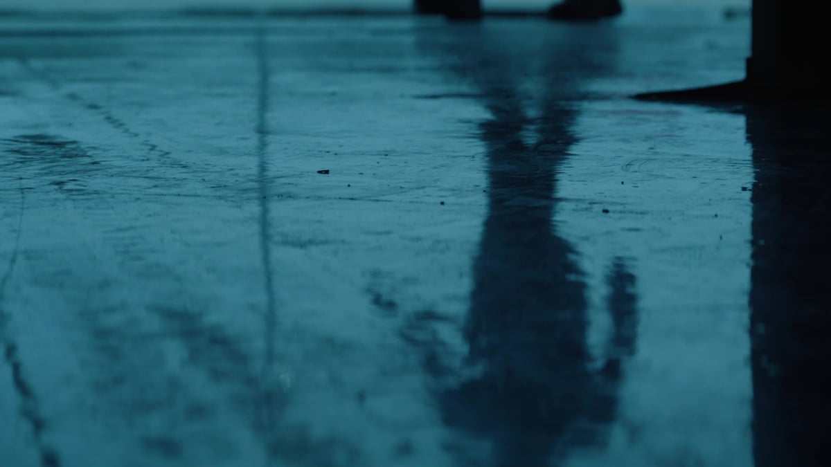 A shadowy silhouette of a person is reflected on a rain-soaked pavement, bathed in a cold blue tone.