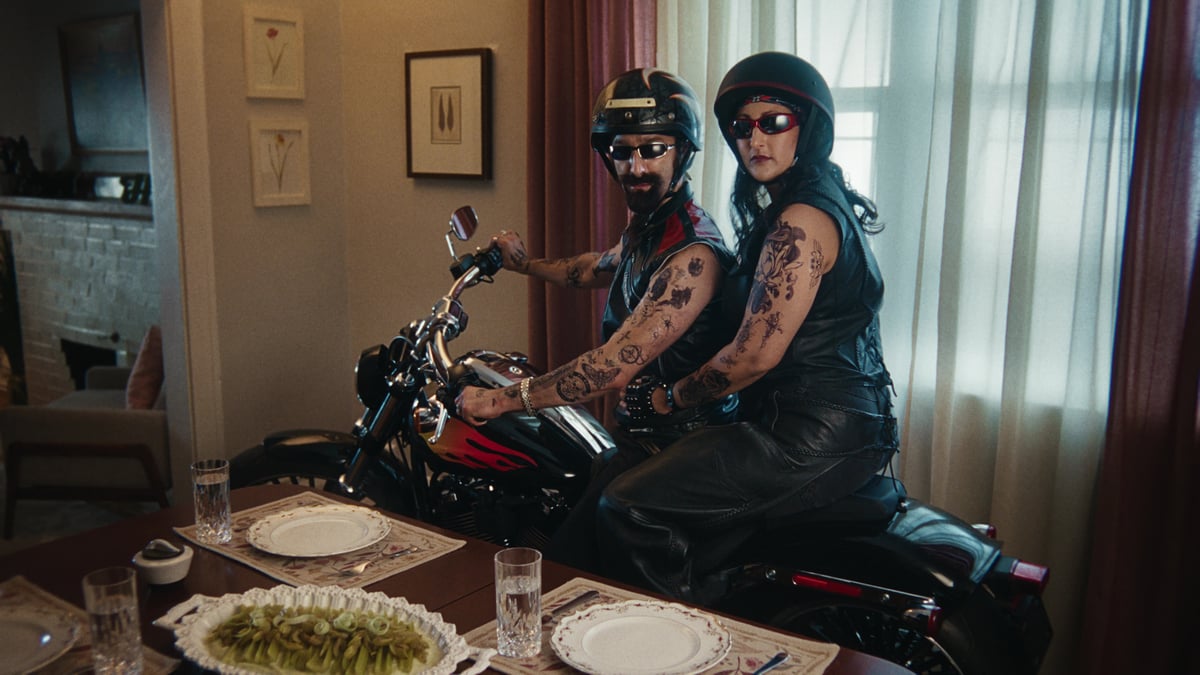 The couple suddenly rides a large motorcycle next to the dining table, dressed like rockstar bikers.