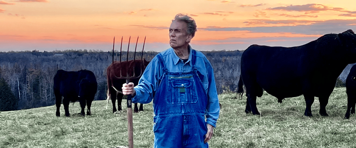 An image of a man holding a farm rake against the backdrop of a beautiful farm with grazing cows.