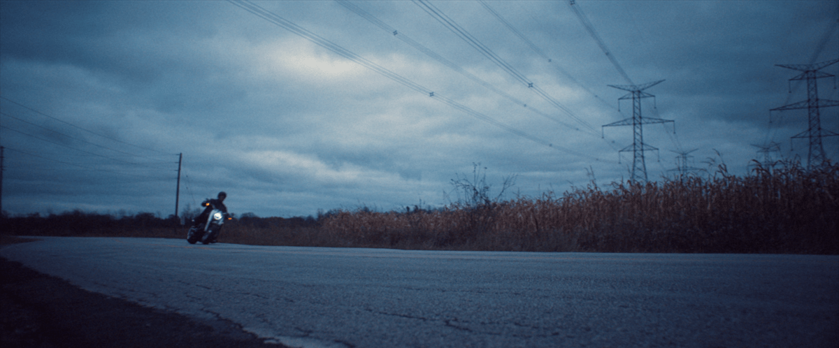A motorcycle approaches from a distance on a road flanked by grass, set against a backdrop of a beautiful sky.