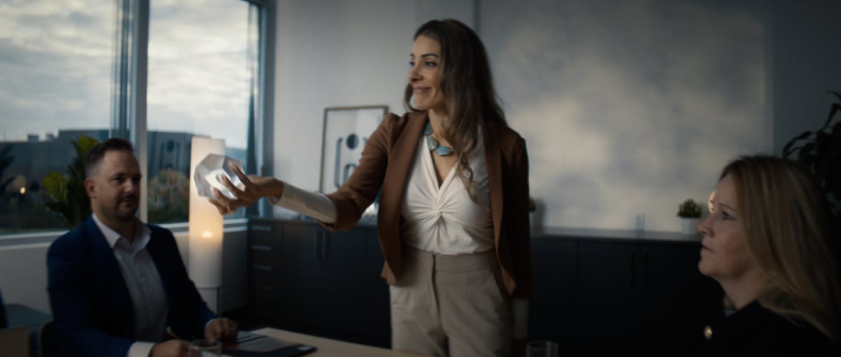 A woman stands before the team, presenting a crystal she holds, while showcasing it to the rest of the team.