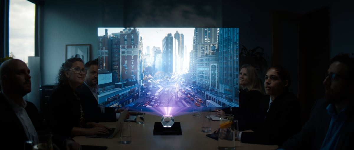 People in a meeting are seated around a table, silently watching a hologram presentation.