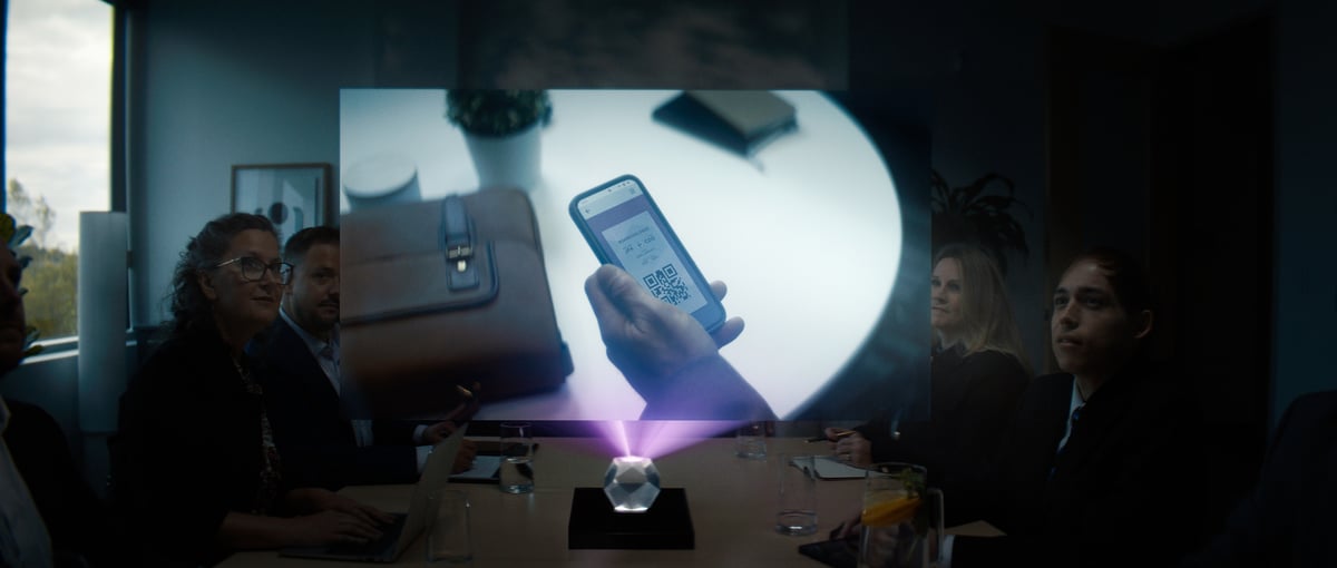  People in a meeting are seated around a table, observing a hologram presentation featuring a hand holding a mobile device.