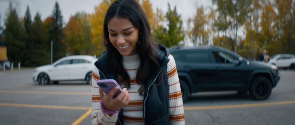A joyful young woman in a parking lot, smiling as she looks at her cellphone.