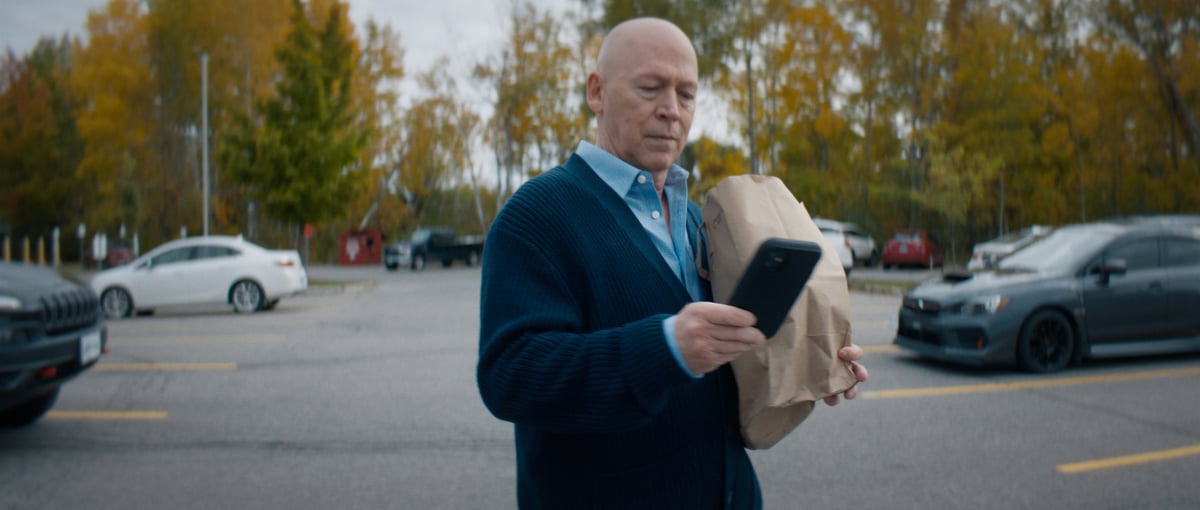 An older man walks in the parking lot, carrying a paper bag with groceries while checking his phone.