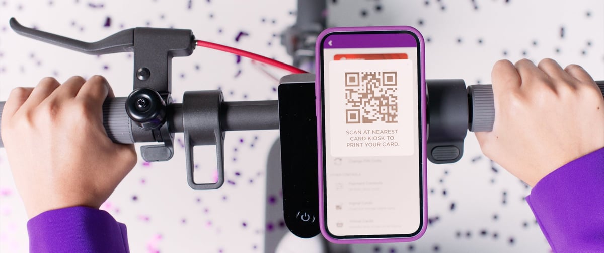 The woman's hand firmly grips the scooter's handlebars, holding a mobile phone with a QR code attached, ready to start the ride.