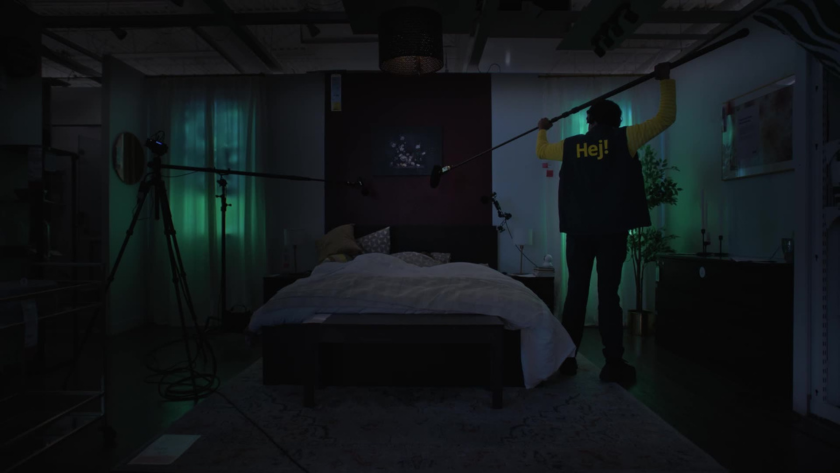 A boom operator captures ambient audio in a dimly lit IKEA bedroom set, preparing for a night of unscripted sleep talk.
