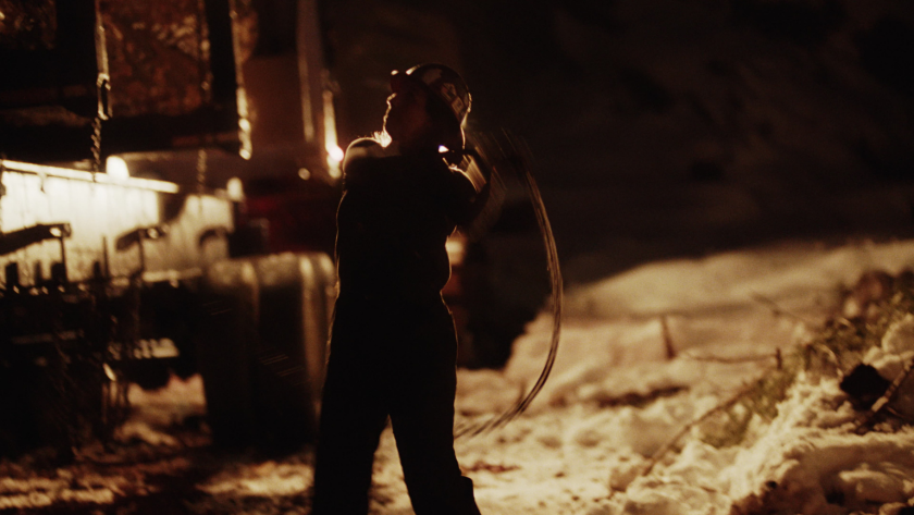 A silhouetted figure swings a chain under dramatic lighting beside a snow-covered truck, capturing the intensity of night operations.