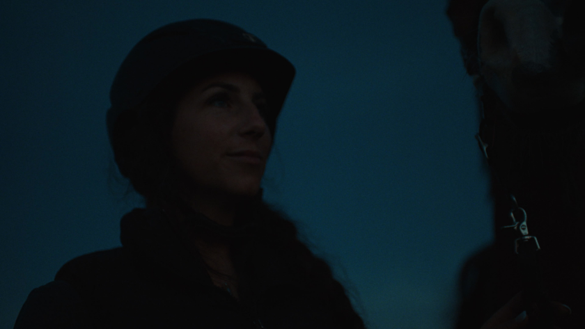 In deep twilight, the driver stands beside her horse, captured in a near-silhouette that mirrors the stillness before the chaos.