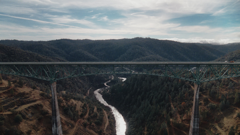 Wide landscape shot of the bridge spanning deep forest valleys under an expansive sky, anchoring the episode’s sense of scale and terrain.