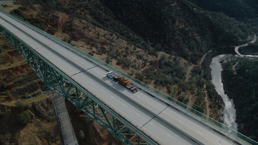 Aerial shot of a truck hauling a dozer across a towering green bridge, emphasizing the vast journey and remote locations.
