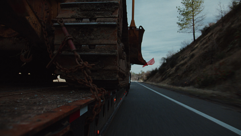 Chains rattle as a massive piece of machinery rolls down the highway, shot from a low angle to intensify movement and power.