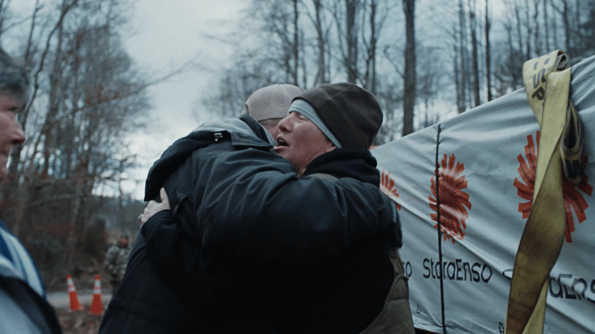 Two people share an emotional hug in front of wrapped lumber with an American flag in the background.