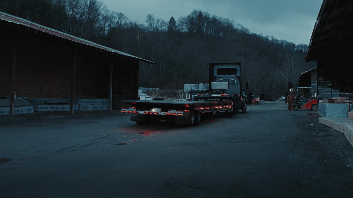 The truck pulls away between two long sheds, its trailer empty but mission in motion, surrounded by soft early light.