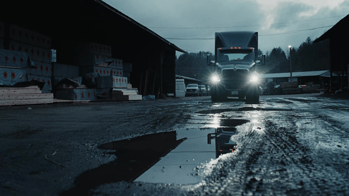 Headlights reflect off a puddle in a muddy lumber yard, framing the truck’s arrival through gritty realism.