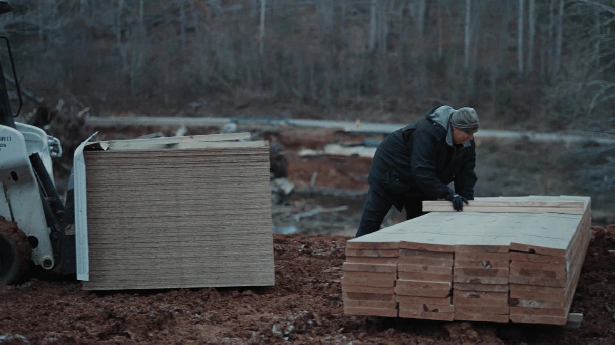 A man stacks and organizes wood at a muddy construction site surrounded by forest.