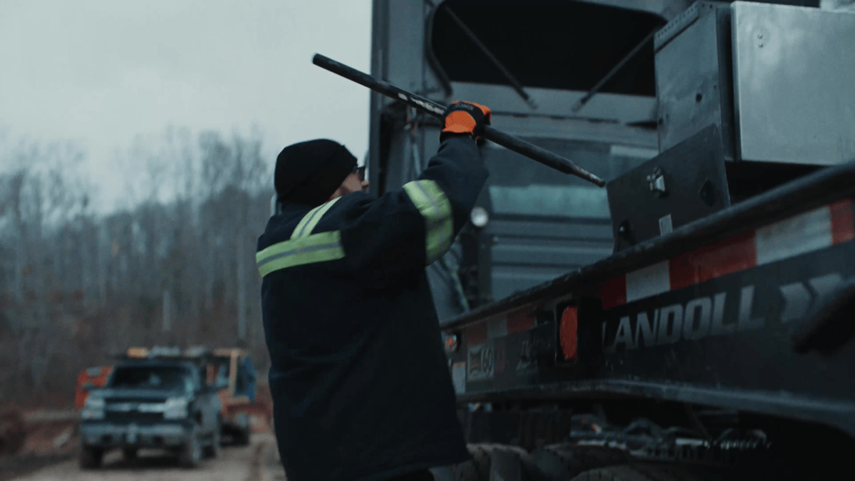 The driver tightens a heavy-duty chain on the trailer in a forest clearing, preparing for another delivery into rugged terrain.