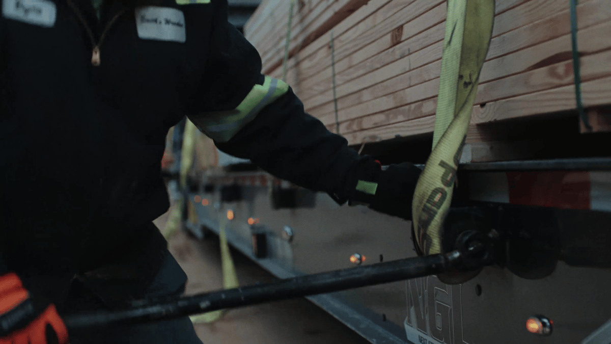 A worker tightens a cargo strap around lumber on the side of a truck trailer.