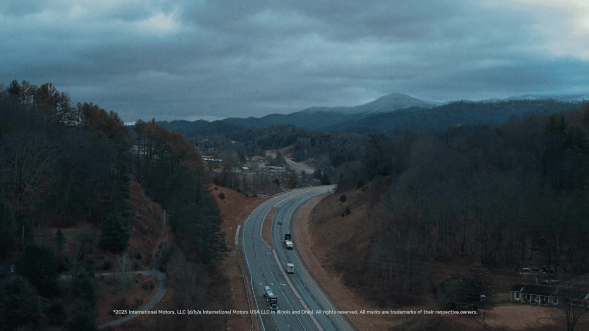 A wide aerial view of a highway winding through forested hills under a cloudy sky.