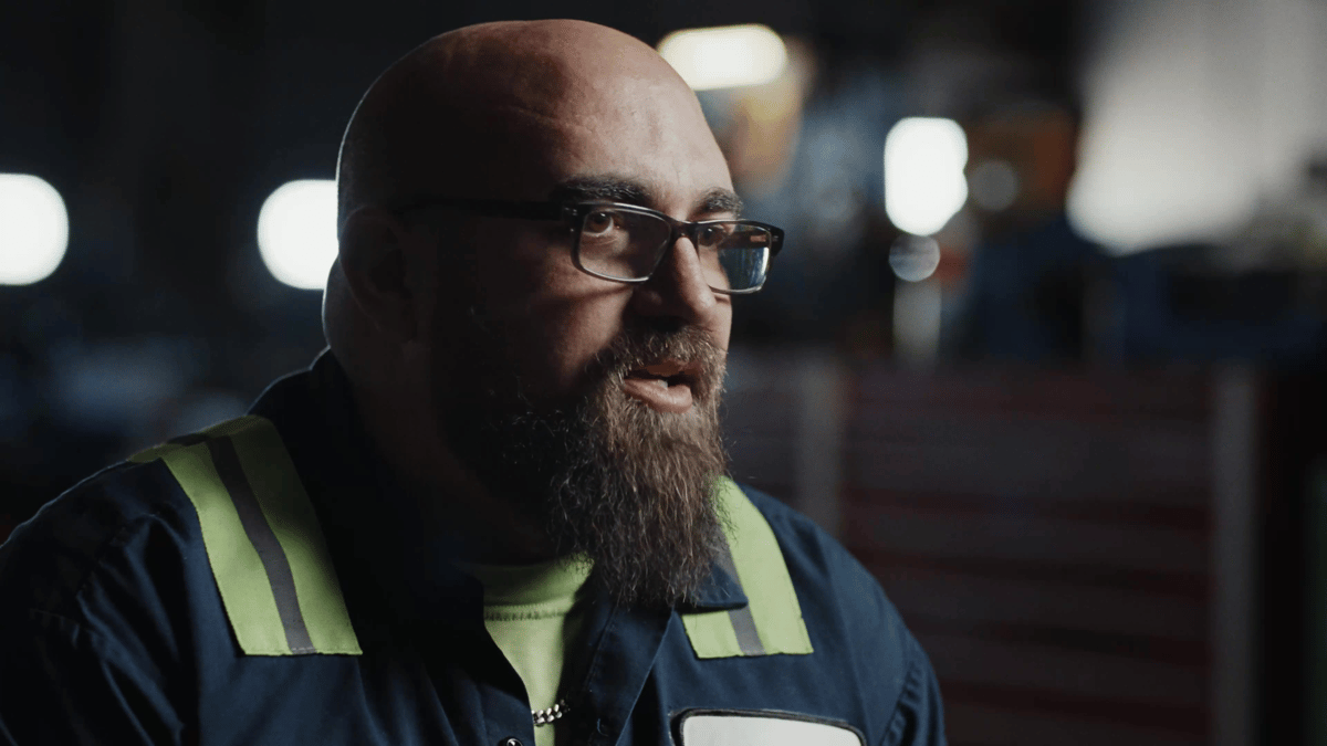 A close-up of a bearded man in reflective workwear speaking during an interview indoors.