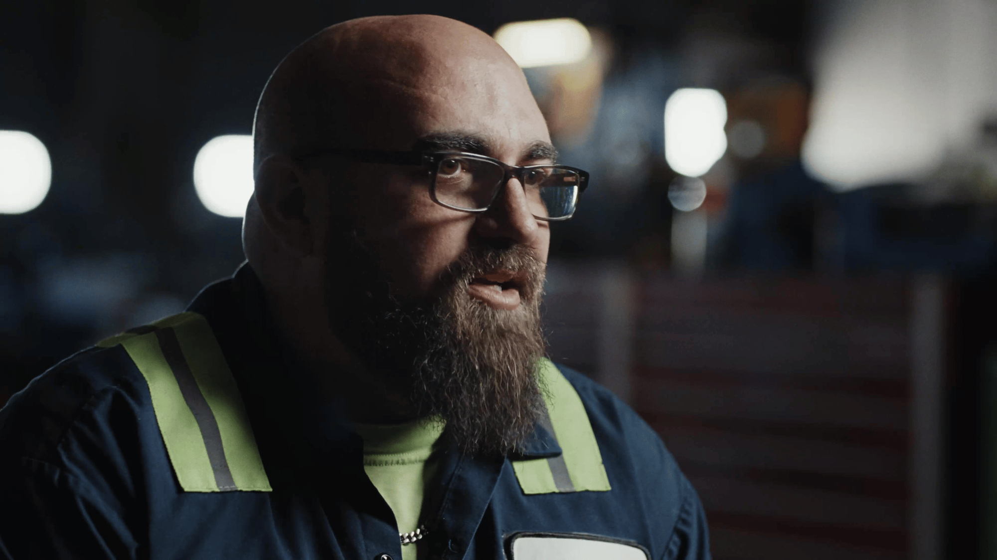 A close-up of a bearded man in reflective workwear speaking during an interview indoors.