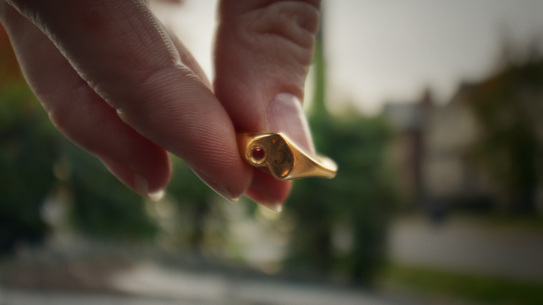 A close-up of a heart-shaped ring held between two fingertips.