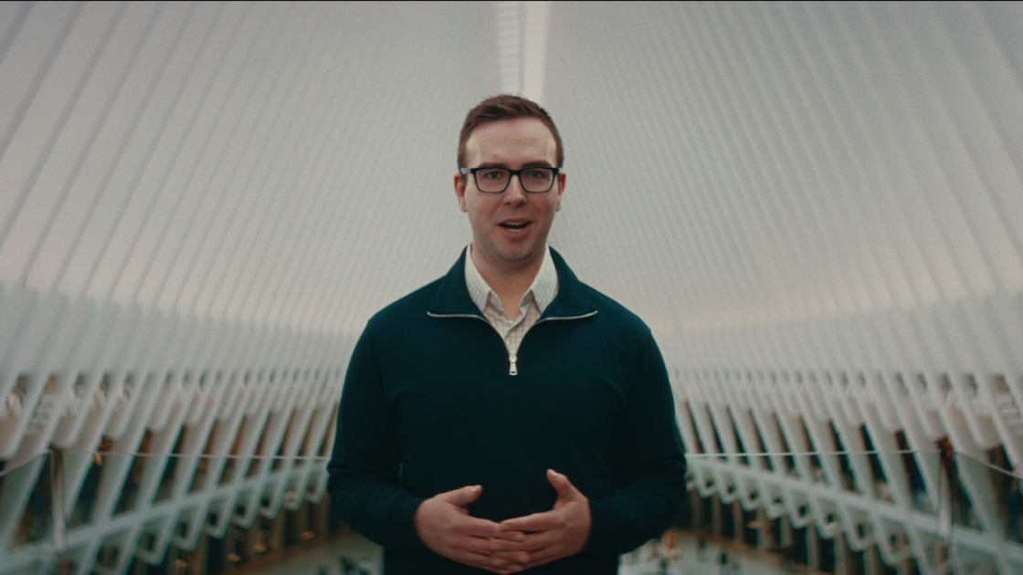 A man in a navy quarter-zip and glasses speaks to camera inside the white ribbed architecture of the Oculus transit hub.