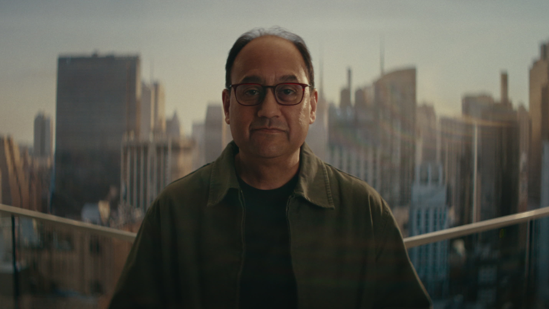 A man with glasses in an olive jacket stands on a rooftop or balcony with a hazy, golden-hour cityscape of skyscrapers stretching behind him.