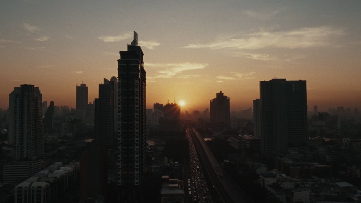A hazy aerial cityscape at sunrise with skyscrapers silhouetted against a golden sky and a highway curving into the horizon.