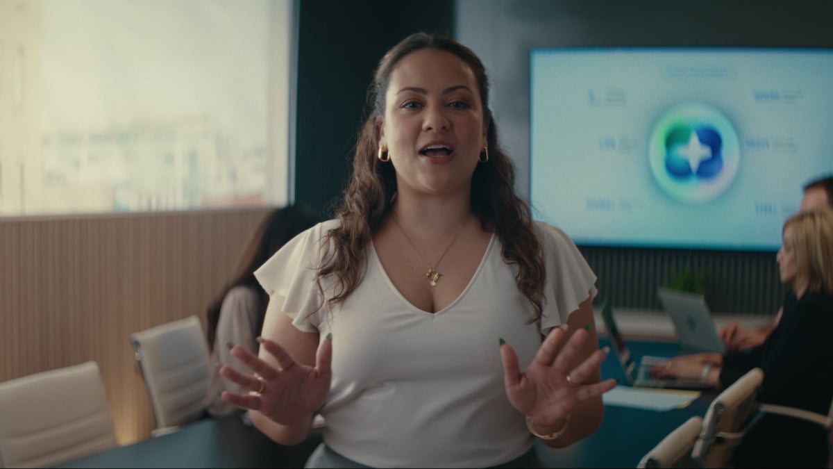 A woman with curly hair in a white v-neck top speaks expressively with both hands raised in a modern conference room, with colleagues working on laptops and a presentation screen behind her.