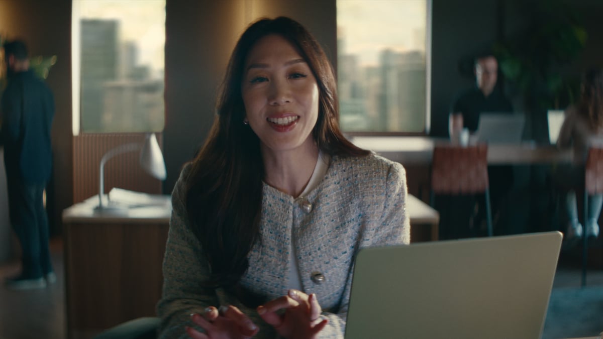 A smiling woman with long dark hair in a textured tweed jacket sits at a laptop in a warmly lit office at golden hour, with colleagues working in the background.