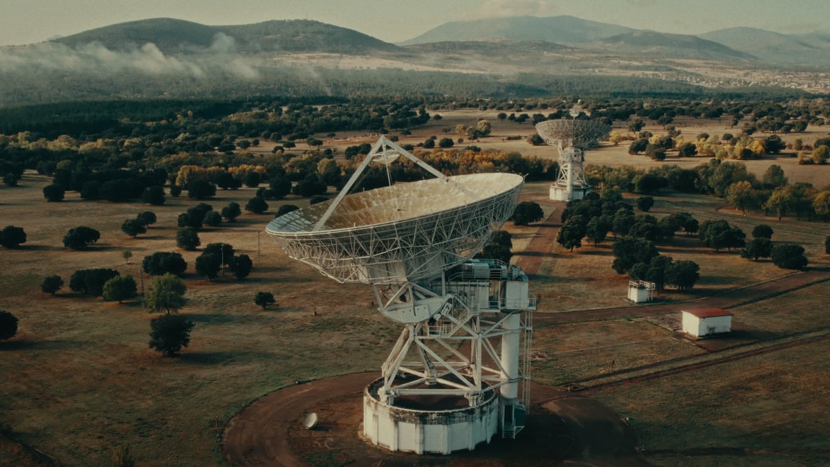 An aerial view of two large white satellite dishes set in a tree-dotted countryside with misty mountains in the distance.